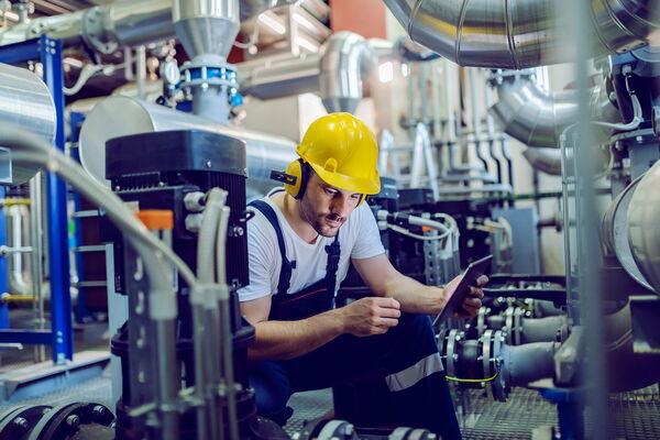In der Serienfertigung dokumentiert eine kniende Person mit Schutzhelm per Tablet einen Arbeitsschritt an Industrieanlagen.